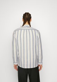 Camisa abotonada de rayas azul claro y blancas, con mangas largas, cuello y dobladillo redondeado. La tela parece suave y texturizada.