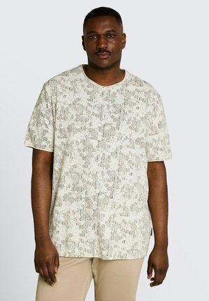 Mężczyzna ubrany w jasnobeżową, wzorzystą koszulę z krótkim rękawem i beżowe spodnie, stojący na tle jednolitego jasnego tła.