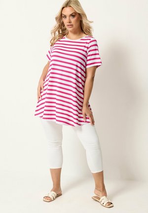 Femme blonde debout portant une tunique ample rayée rose et blanche, des leggings blancs courts et des sandales beige.