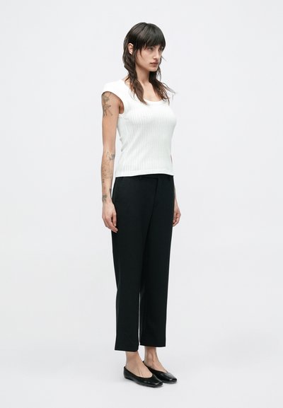 Λευκή ριμπ τοπ χωρίς μανίκια, μαύρο ραμμένο παντελόνι και μαύρα επίπεδα παπούτσια. Το σύνολο χαρακτηρίζεται από εφαρμοστή σιλουέτα με μινιμαλιστικό σχεδιασμό.