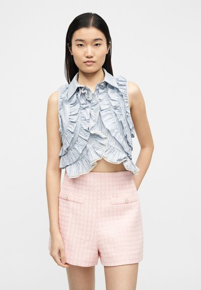 Νεαρή γυναίκα που φοράει μπλε και λευκό ριγέ crop top χωρίς μανίκια με βολάν και ροζ υφασμένα σορτς ψηλόμεσα, στέκεται μπροστά σε απλό φόντο.