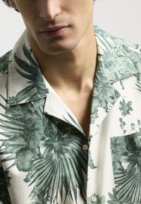 Camisa de manga corta con un estampado de hojas tropicales verdes sobre un fondo crema, con un bolsillo en el pecho y un diseño de cuello clásico.