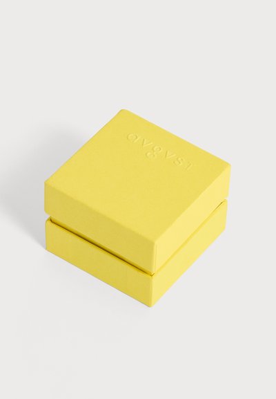 Boîte carrée jaune vif fermée avec texte en relief sur le couvercle, placée sur une surface blanche unie.