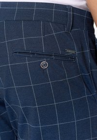 Marineblå ternede bukser med nærbillede af ryggen, der viser en lomme med knap og en lille labeldetalje. Struktureret stof med rutenetmønster.
