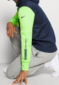 Nike hoodie featuring a navy body and bright green sleeves, with "SEAHAWKS FOOTBALL" text on the left arm. Gray sweatpants and white sneakers.