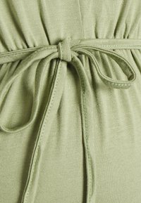 Tecido verde claro com uma textura suave, apresentando um design de cintura amarrada e detalhes de costura ao longo das bordas.