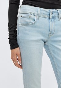 Τζιν παντελόνι από ανοιχτό μπλε φθαρμένο denim με στενή εφαρμογή, διαθέτει πέντε τσέπες, μεταλλικά κουμπιά και ομαλή υφή. Συνοδεύεται από ένα μαύρο μπλουζάκι με μακριά μανίκια.