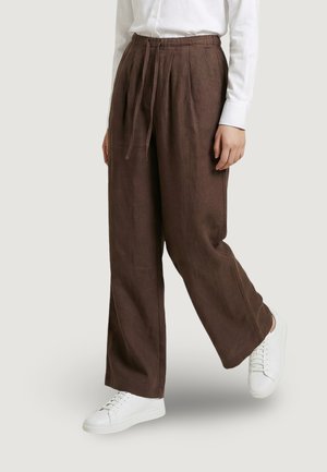 Persona che indossa pantaloni marroni larghi e larghi con vita regolabile, camicia bianca abbottonata e sneakers bianche, in piedi su uno sfondo neutro.