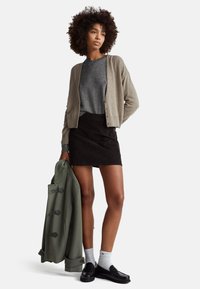 Młoda kobieta o kręconych włosach, ubrana w szarą bluzkę, beżowy kardigan, czarną spódnicę, białe skarpetki i czarne mokasyny, trzymająca w ręku zielony płaszcz, stoi.