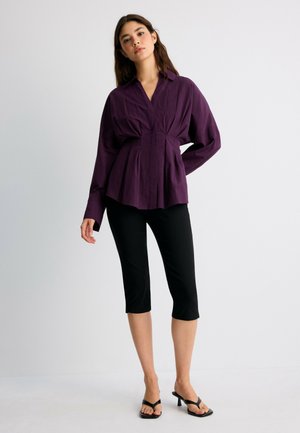 Jeune femme portant un chemisier violet foncé à manches longues, un pantalon court noir et des sandales à talons noires sur un fond clair uni.