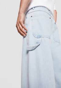 Pantaloni larghi in denimazzurro chiaro con grandi tasche posteriori, un dettaglio con patta laterale e accenti di cucitura. Texture liscia e vestibilità rilassata.