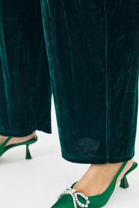 Fluwelen donkergroene wijde broek gecombineerd met groene haksandalen met een strass strikaccent. Glad materiaal en een slank ontwerp.