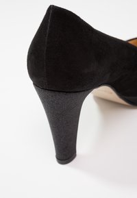Chaussure à talon haut en suède noir avec un bout pointu et un talon noir texturé. Le talon est fin et présente une finition brillante.