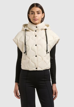 Młoda kobieta ubrana w bezrękawnik pikowany w kolorze kremowym z wysokim kołnierzem i czarnymi sznureczkami, na czarnej bluzce z długim rękawem i spodniach.