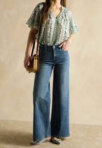 Blouse blanche à motifs avec des détails floraux verts, jean taille haute bleu clair et chaussures plates. Petit sac à bandoulière beige.