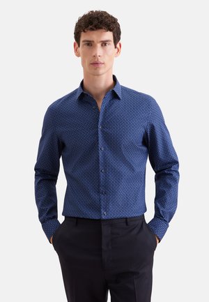 Mann mit lockigem Haar, der ein eng anliegendes blau gemustertes Langarmhemd und schwarze Hose trägt, steht mit den Händen in den Taschen.