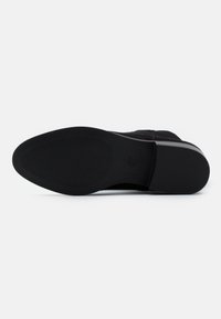 Suela de zapato negra con goma texturizada para agarre, con un talón liso y una forma general redondeada. Diseño minimalista, sin patrones visibles.