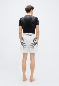 Μαύρο κοντομάνικο ρούχο προστασίας από την ηλιακή ακτινοβολία, σε συνδυασμό με λευκά σορτς που έχουν μαύρα φυλετικά μοτίβα και λογότυπο «VENUM» στη μέση.