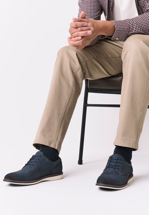 Muž sediaci na stoličke, oblečený v béžových nohaviciach, tmavomodrých ponožkách a tmavomodrých šnurovacích topánkach s bielymi podrážkami, ruky zložené a odpočívajúce na kolene.