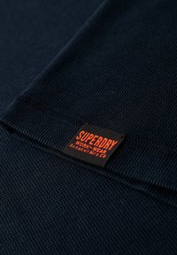 Close-up of navy blue textured fabric with a black label reading "SUPERDRY WORK-WEAR GARMENT MFG CO" in orange text.