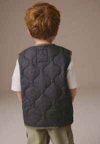 Vestă matlasată bleumarin cu un model ondulat, margini verzi și design fără mâneci, purtată peste o cămașă albă. Vedere din spate care evidențiază textura și forma.