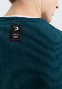 Türkiser strukturierter Strickpullover mit einem schwarzen Markenetikett auf der Rückseite, das gerippte Muster und ein Rundhalsausschnitt-Design hervorhebt.