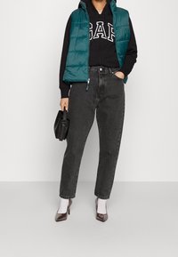 Turkos vadderad väst över en svart hoodie med vit text, i kombination med hög midja svarta jeans och glänsande bruna högklackade skor. Svart handväska.