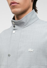 Veste grise avec un col haut, boutons-pression métalliques et un tissu texturé. Présente un logo brodé blanc sur la poitrine.