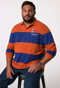 Langärmliges Polo-Shirt mit orange-blauen Streifen und gesticktem Design "Ocean Adventure". Getragen mit Jeans, sitzt auf einem schwarzen Hocker.