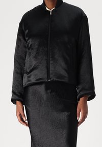 Svart kort bomberjacka med en slät, strukturerad yta, dragkedjestängning och sidoficka. Matchar med en koordinerad kjol.