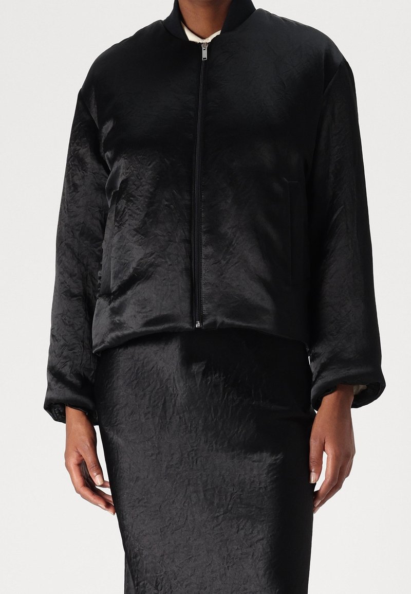 Svart kort bomberjacka med en slät, strukturerad yta, dragkedjestängning och sidoficka. Matchar med en koordinerad kjol.