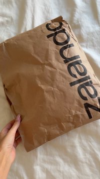 Mano che tiene un pacchetto di carta marrone stropicciato, con del testo nero parzialmente visibile su una superficie di tessuto bianco.