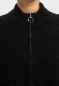 Juodas megztinis su užtrauktuku, aukštu apykakliu, sidabrinės spalvos užtrauktuko detalėmis ir tekstūruota, pūkuota medžiaga. Matomų raštų ar puošmenų nėra.