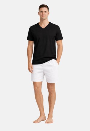 Muž stojící naboso, oblečený v černém trikotu s výstřihem do V a bílých šortkách, s jednou rukou v kapse na jednobarevném světlejším pozadí.