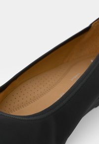 Scarpa piatta nera con punta tonda. Presenta un'esterno liscio e una soletta imbottita di colore marrone chiaro con perforazioni per la ventilazione.