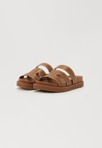 Brune sandaler laget av syntetisk materiale med brede, kryssende reimer med hvite sømmer og en tykk, teksturert såle.