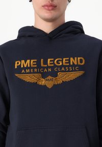 Marineblauwe hoodie met een kangoeroezak, versierd met gouden geborduurde tekst "PME LEGEND AMERICAN CLASSIC" en een vleugelontwerp.