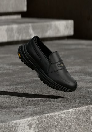 Mustat nahkaiset slip-on-kengät, joissa on karkeapohja, kelluvat betonisten portaiden yläpuolella pehmeässä valaistuksessa.