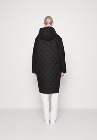 Abrigo largo acolchado negro con capucha, con un patrón de diamantes y hombros caídos, llevado sobre pantalones blancos y zapatillas.