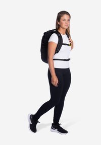 Zwarte rugzak met verstelbare banden, gedragen door een persoon in een wit shirt en zwarte leggings. Kenmerkt zich door een minimalistisch ontwerp en meerdere gespen.