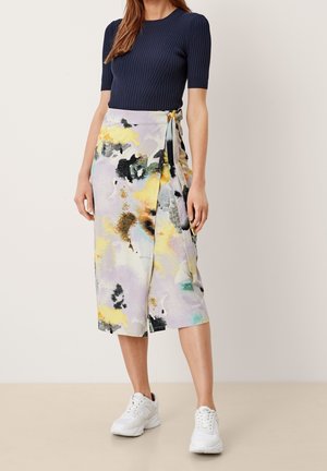 Nainen, joka on pukeutunut laivastonsiniseen, lyhythihaiseen, ribbattuun toppiin, pastellinsävyiseen abstraktikuvioiseen midimittaisen kietaisuhameeseen ja valkoisiin tennareihin, seisoo yksinkertaista taustaa vasten.