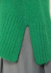 Πράσινο πλεκτό πουλόβερ με ριχτή υφή και λεπτομέρεια σε V στο στρίφωμα, συνδυασμένο με ένα γκρίζο ρούχο από κάτω. Μαλακό, παχύ ύφασμα.