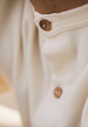 Camisa de color beige claro con cuello que presenta un detalle de botón de madera en el cuello, fabricada en una suave mezcla de algodón con una textura lisa.