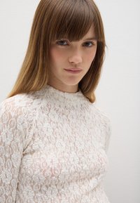 Top bianco in pizzo con scollatura alta, maniche lunghe e motivi floreali; texture delicata e finitura semitrasparente.