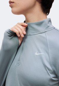 Άτομο που φοράει μπλουζάκι Nike ανοιχτό μπλε με φερμουάρ μέχρι το στήθος, τραβά το φερμουάρ κοντά στον λαιμό, με ορατό το λογότυπο Nike στο στήθος.