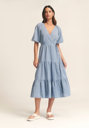 Blue and white checked midi dress with a V-neckline, short flutter sleeves, tiered ruffle hem, and a drawstring waist. Worn with sandals.