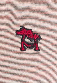 Animal stylisé brodé rouge et noir avec une queue relevée sur un tissu blanc à fines rayures horizontales rouges.