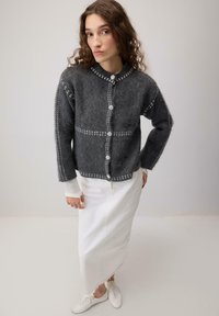 Ženska s kodrasto lase, oblečena v temno sivo suknjič z gumbi, dolgo belo krilo in bele čevlje, ki stoji ob enotnem svetlem ozadju.