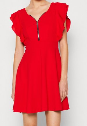 Femme portant une robe rouge vif avec des manches à volants et une fermeture éclair à l’avant partiellement ouverte, debout devant un fond clair uni.