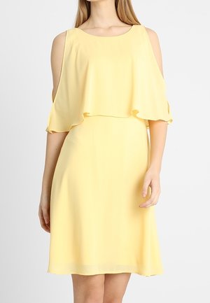 Kvinde iført en gul kjole uden ærmer, der går til knæene, med udskæringer ved skuldrene og en lagdelt, flydende overdel.